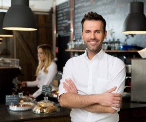 Successful small business owner standing with crossed arms with employee in background preparing coffee